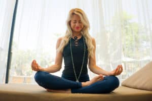 Eine Frau meditiert in einer Lotusposition mit geschlossenen Augen in einer ruhigen Innenumgebung und trägt Mala-Ketten aus Heilsteinen.