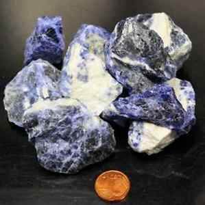 Eine Ansammlung roher blauer und weißer Sodalithsteine wird auf einer dunklen Oberfläche mit einem US-Penny als Größenvergleich präsentiert.