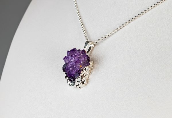 Vor einem weißen Hintergrund ist eine silberne Halskette mit einem Anhänger mit einem groben violetten Edelstein zu sehen.