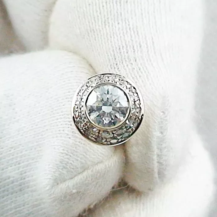 Eine behandschuhte Hand hält einen runden Diamantring mit einem großen Mittelstein, der von einem Kranz kleinerer Diamanten umgeben ist, die in ein Metallband eingefasst sind.