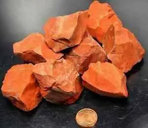 Haufen unregelmäßig geformter roter Steine neben einer Kupfermünze zum Größenvergleich, auf einer schwarzen Oberfläche platziert.
