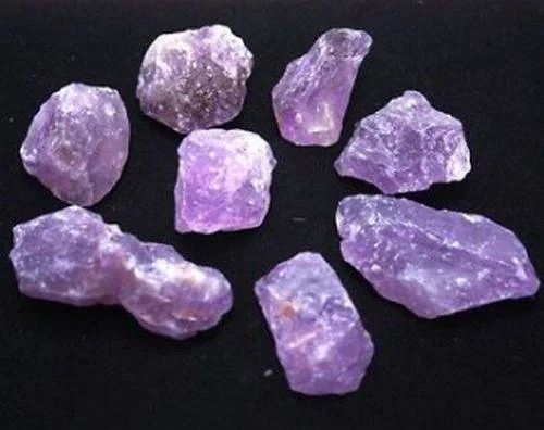 Acht rohe, ungeschliffene violette Kristalle sind auf einer schwarzen Oberfläche verteilt.