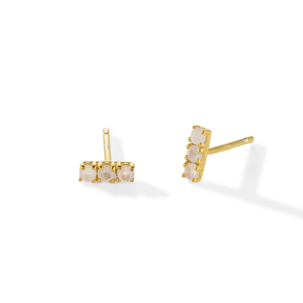 Ein Paar goldene Ohrstecker mit einer horizontalen Reihe aus drei kleinen weißen Steinen auf weißem Hintergrund.